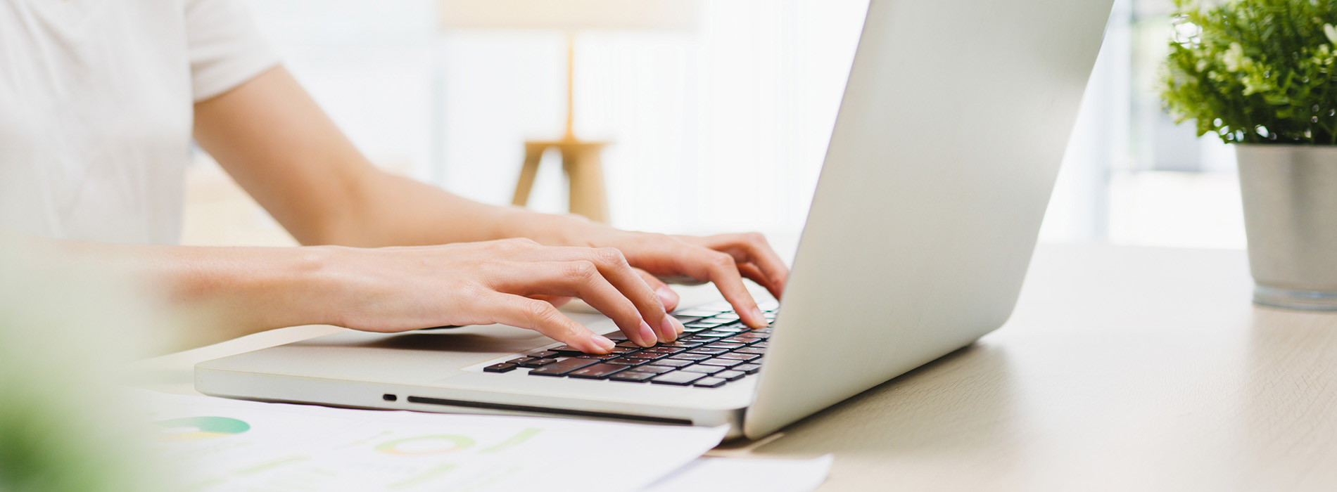 The image shows a person typing on a laptop at a desk with a blurred background.